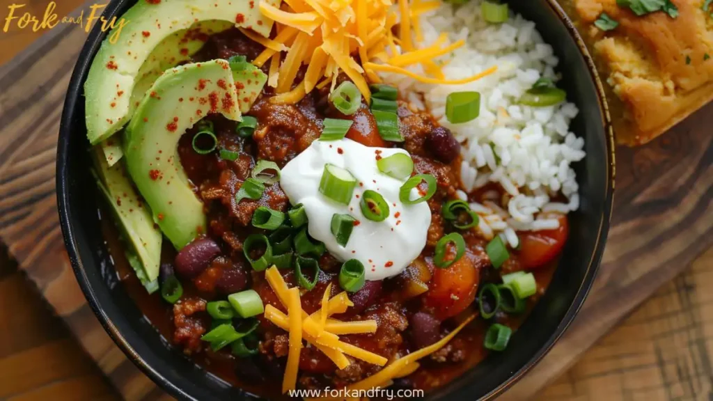 Overhead shot of venison chili garnished with cheddar cheese, sour cream, avocado, and green onions, served with cornbread.