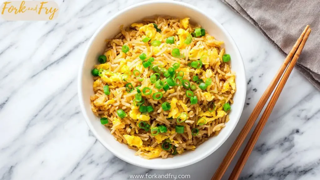 Overhead view of egg fried rice in a white bowl, topped with chopped green onions, with wooden chopsticks on the side and a marble kitchen counter background.